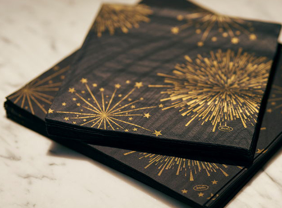 Stack of festive black napkins with gold fireworks and starburst patterns on a marble surface.