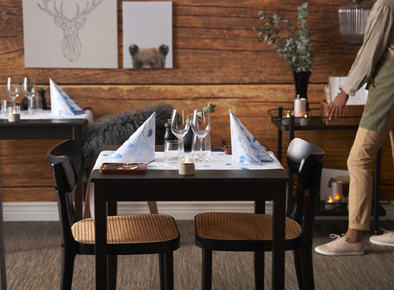 Rustic dining setup with neatly arranged tables, woven chairs and soft candlelight – set against wood panelled walls with blue and winter tableware.