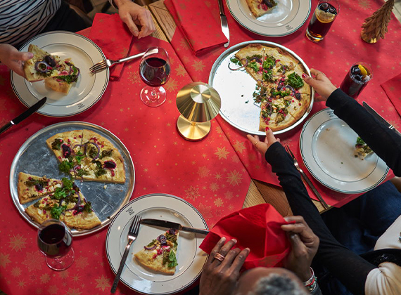 Festive table setting with pizzas, wine and soda, surrounded by diners reaching for slices – evoking warmth and togetherness.