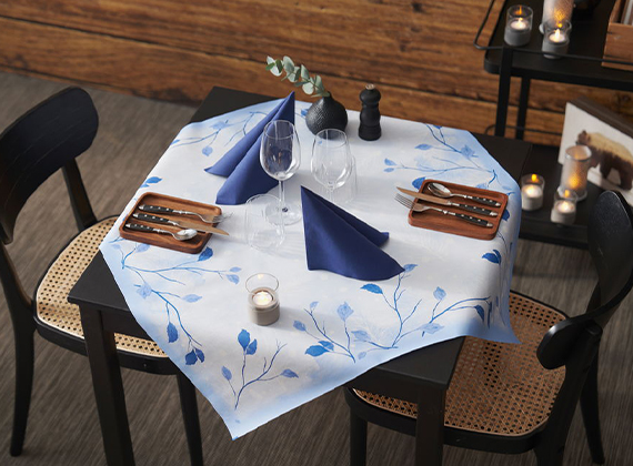 Intimate festive table setting for two with blue leaf-patterned tablecloth, wooden trays, folded blue napkins and tealight candles. Perfect for cosy holiday dining décor.