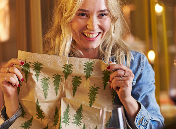 Woman holding a festive napkin with Christmas tree print at a dining table.
