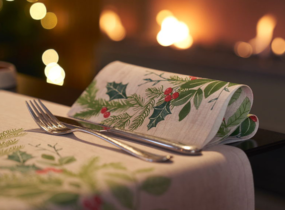 Festive holiday dining table with holly-themed napkin, cutlery neatly placed and warm fireplace glowing in the background.