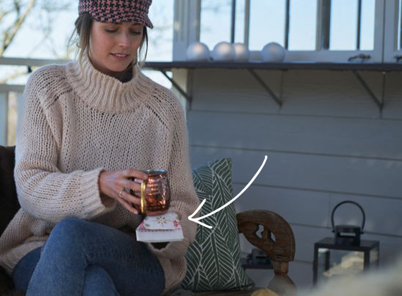 Outdoor winter setting with two people on a porch – one seated on a fur-covered bench holding a hot beverage with a festive Nordic-style napkin.
