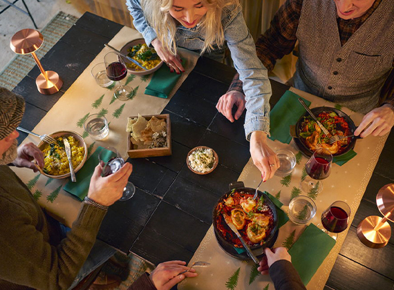 Group of four enjoying a festive meal around a rectangular table set with green napkins, red wine and pine tree-themed table décor, capturing a warm communal dining experience.