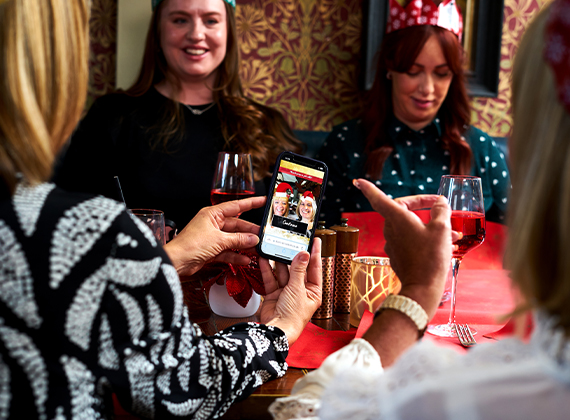 Group of people wearing festive paper crows seated around a table, sharing content on their smartphones during a holiday gathering.